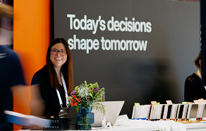 Smiling woman standing behind a booth with a sign reading 'Today's decisions shape tomorrow', flowers and brochures on the counter.