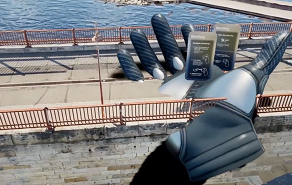 Giant metallic hand sculpture extending over a stone seawall by the water, with railings and a boardwalk in the background.