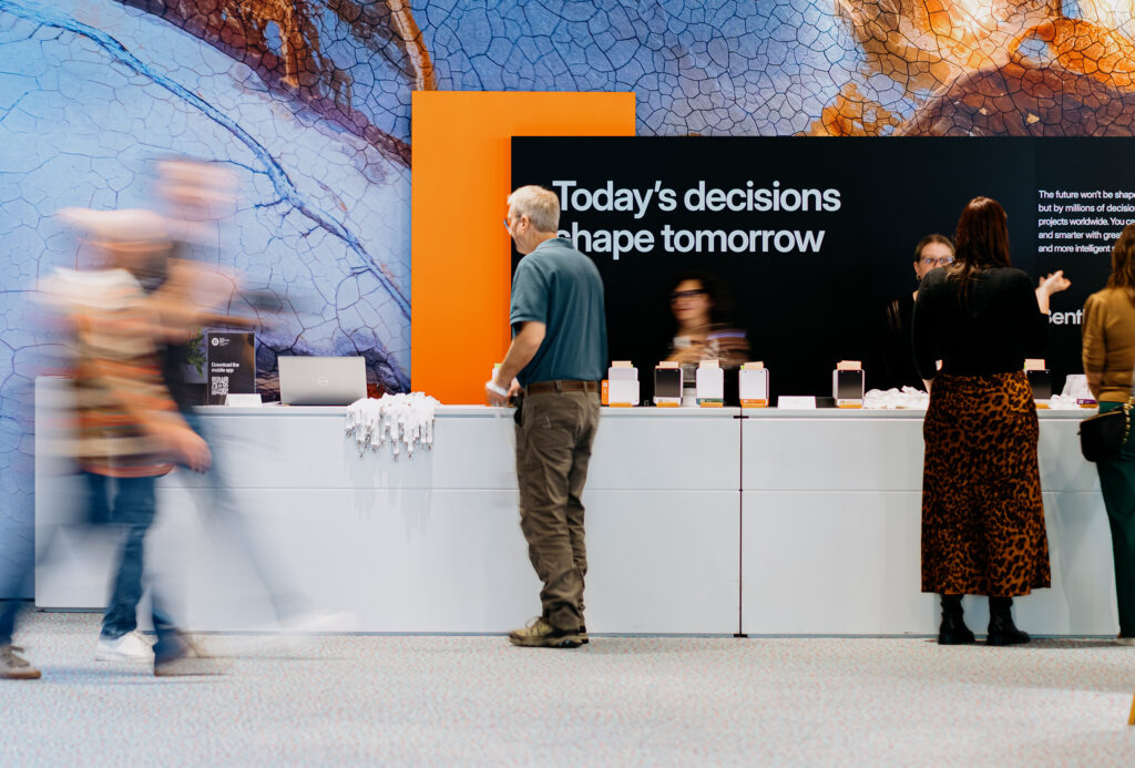 Exhibition booth with a black wall displaying the slogan “Today’s decisions shape tomorrow” and an orange frame, shoppers browsing devices on a counter behind them.