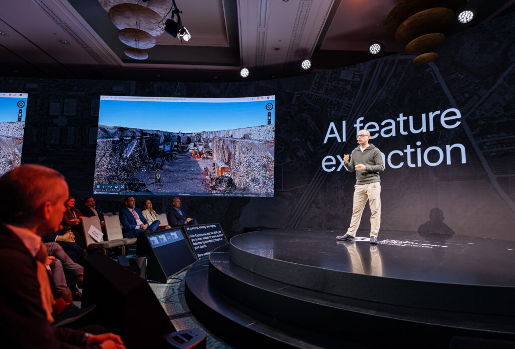 Patrick Cozzi, founder of Cesium, on stage at a tech talk about "AI feature extraction," with a digital twin of a construction site displayed on the screen behind him.