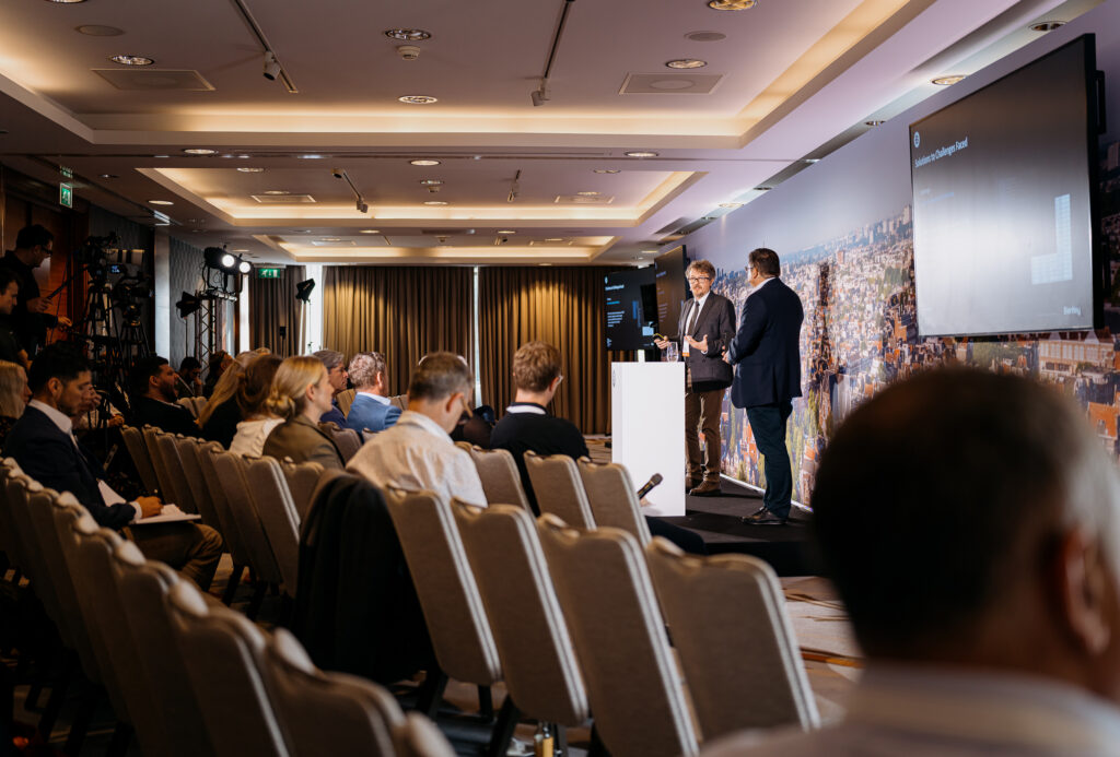 Two men in suits speak on a stage during a conference, with a large cityscape backdrop and an attentive audience in rows of seats.