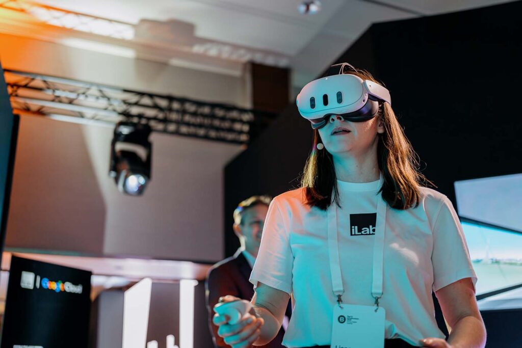 Woman wearing a VR headset at a technology demo, badge visible, reaching toward something off-screen in a conference setting