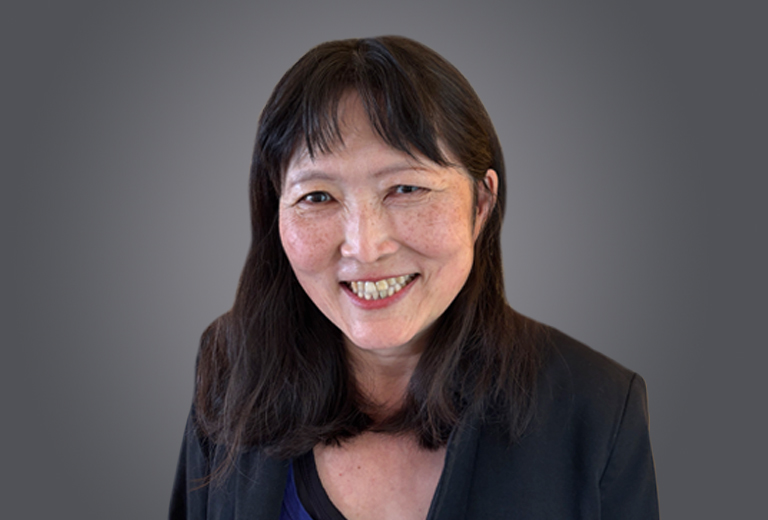 Smiling woman with dark hair in a black blazer against a gray gradient backdrop, looking at the camera.