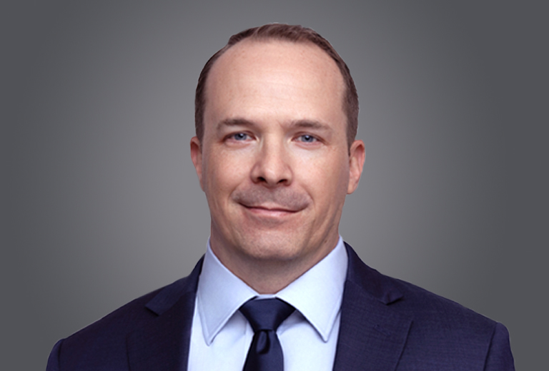 Professional headshot of a smiling man in a dark suit and navy tie against a gray gradient background.