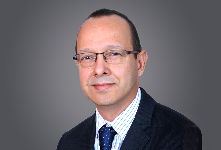 Professional headshot: man in a navy suit, striped shirt, blue tie, and glasses against a gray backdrop.