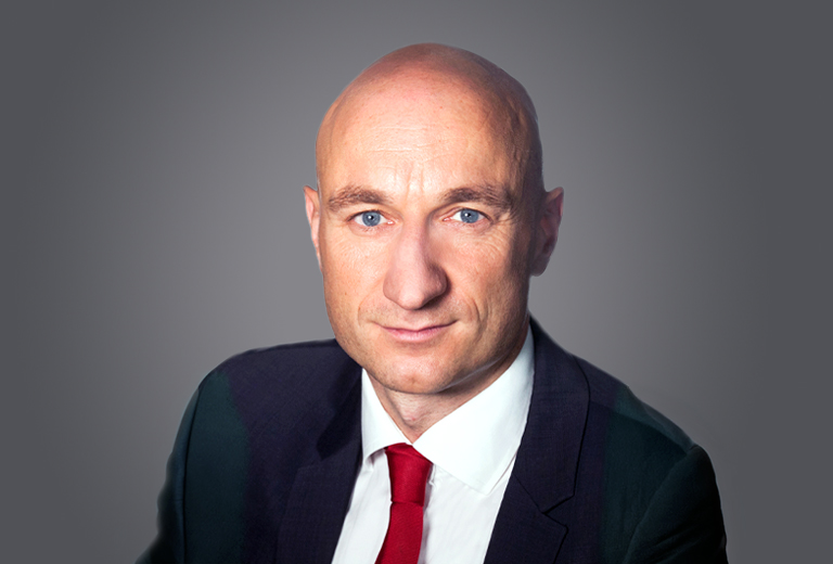 Professional head-and-shoulders portrait of a bald man in a dark suit and red tie, looking at the camera against a neutral gray background.