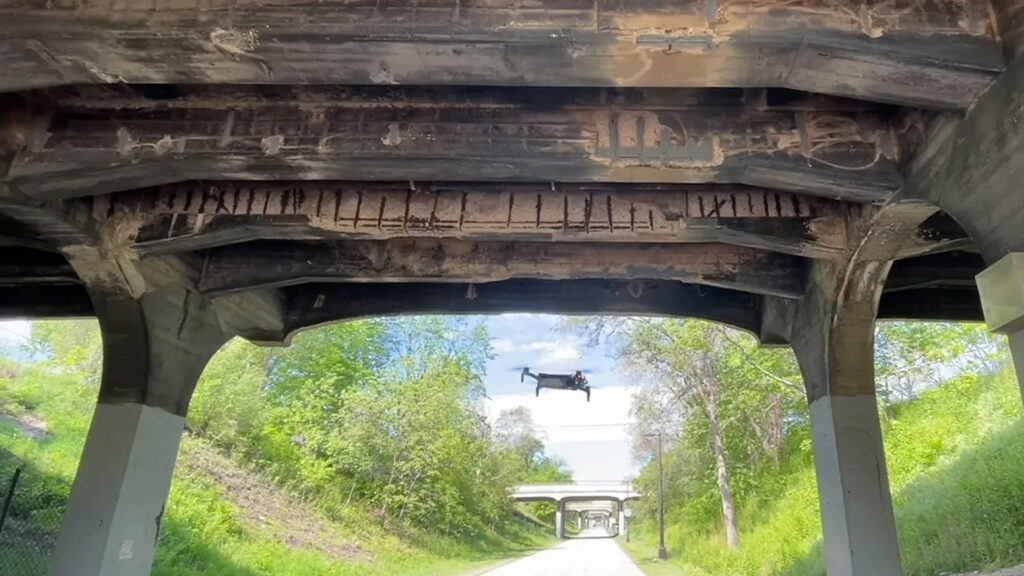Under-pillar view of a weathered wooden bridge with rusted planks; a drone hovers over the road beneath, inspecting the structure