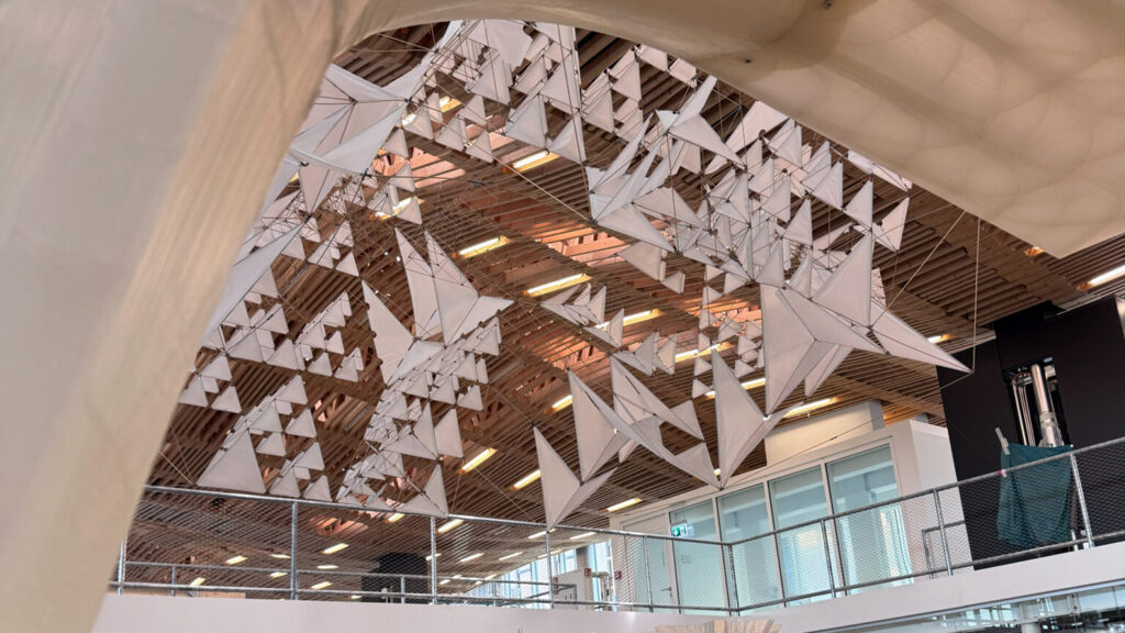 Indoor atrium with a sculptural installation of hanging white triangular sails suspended from a wooden ceiling.