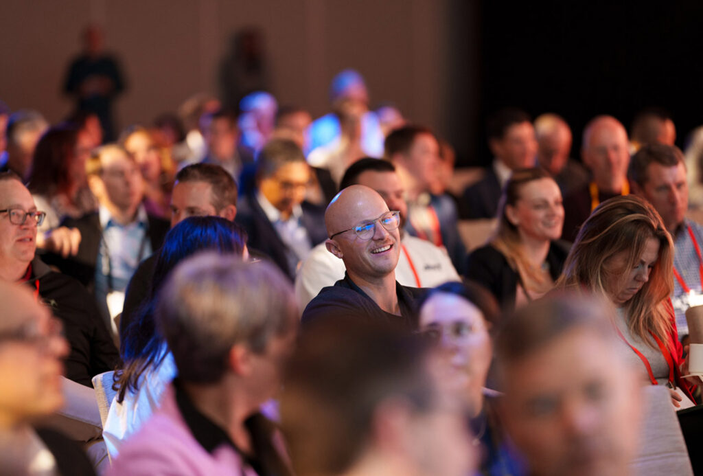 Crowd of people at a conference or event, some smiling, with a man wearing glasses in the foreground center.