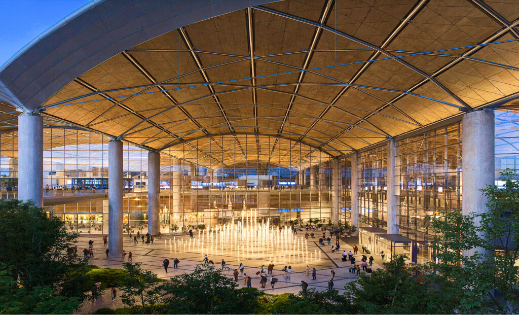 An evening view of a large, modern airport terminal with a high, vaulted ceiling and a central plaza featuring an illuminated fountain, a finalist project by Port Polska.