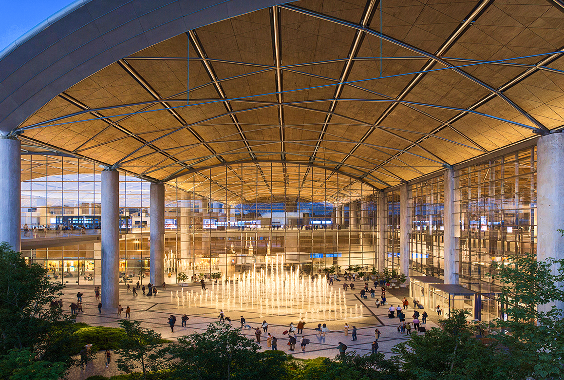 An evening view of a large, modern airport terminal with a high, vaulted ceiling and a central plaza featuring an illuminated fountain, a finalist project by Port Polska.