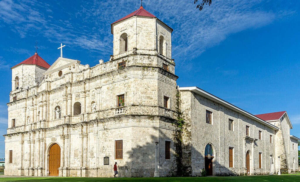 A wide shot of the historic Our Lady of Light Parish Church in Loon, a beautifully restored stone building with twin bell towers against a clear blue sky, a finalist project by E.S. de Luna.