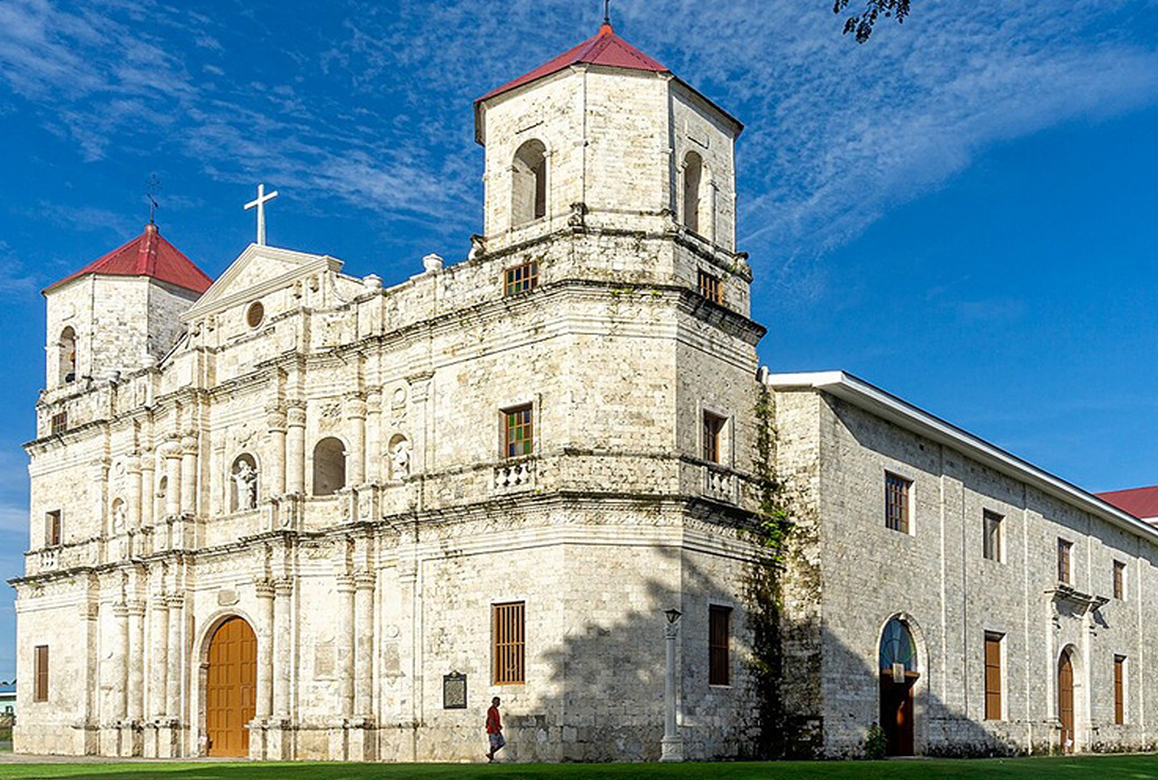 A wide shot of the historic Our Lady of Light Parish Church in Loon, a beautifully restored stone building with twin bell towers against a clear blue sky, a finalist project by E.S. de Luna.
