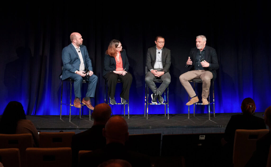 Four people in business attire sit on high chairs on a stage, engaged in a panel discussion, with an audience seated in front of them.