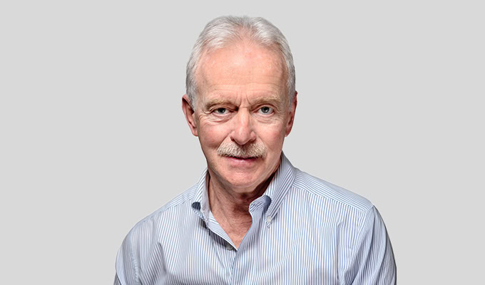 Older man with white hair and mustache, wearing a striped button-up shirt, poses against a plain gray background.