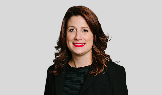 Eine Frau mit schulterlangem braunem Haar, einem schwarzen Blazer und rotem Lippenstift lächelt vor einem schlichten grauen Hintergrund.