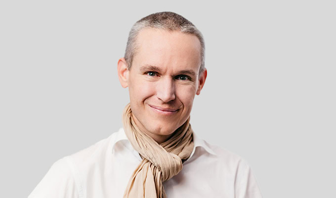 Eine Person mit kurzen grauen Haaren lächelt, trägt ein weißes Hemd und einen beigen Schal vor einem schlichten hellgrauen Hintergrund.