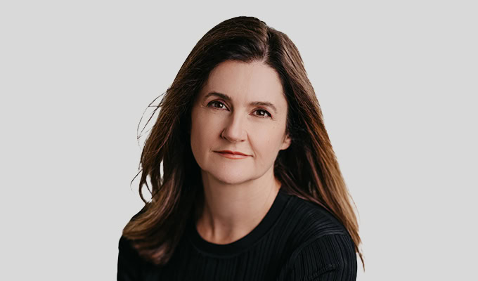 Eine Frau mit langen braunen Haaren und einem schwarzen Oberteil posiert vor einem hellgrauen Hintergrund.