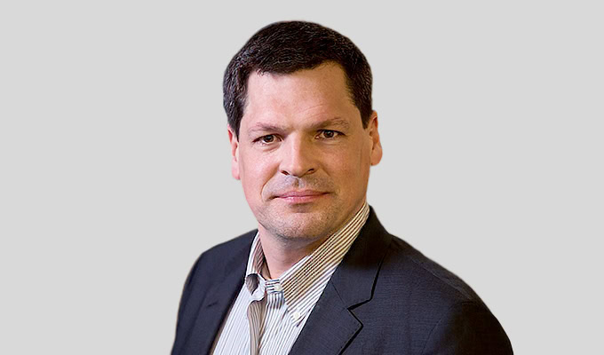 Ein Mann mit kurzen dunklen Haaren, einem dunklen Jackett und einem hell gestreiften Hemd, posiert vor einem schlichten hellgrauen Hintergrund.