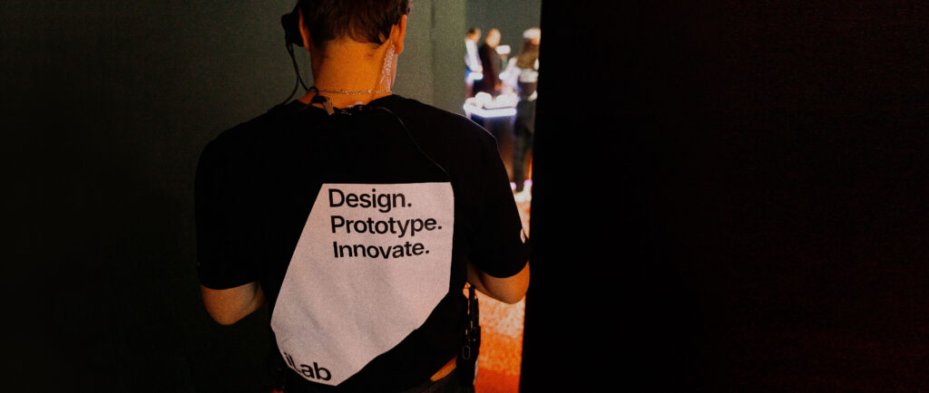 A person wearing a black t-shirt with the words "Design. Prototype. Innovate." stands facing away, partially obscured in a dimly lit environment.