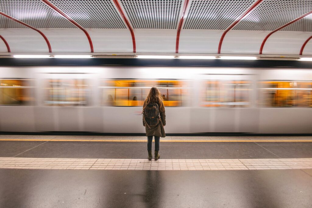 A person wearing a backpack stands on a subway platform as a train passes by in the background.