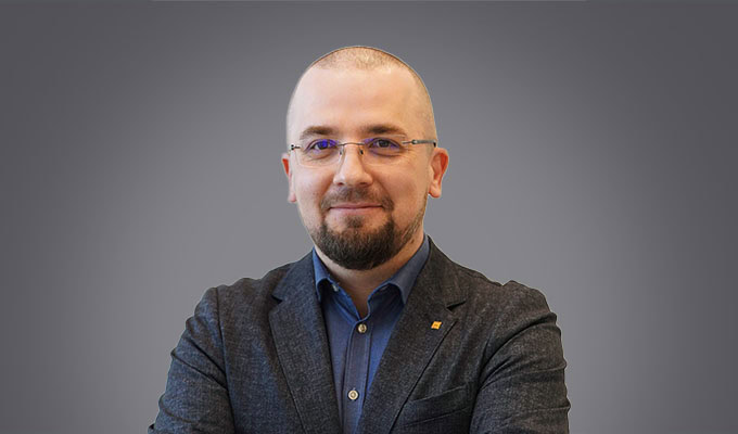 Andrius Jurelionis, a man with a shaved head, glasses, and a beard, stands with arms crossed in a dark blazer and blue shirt against a plain gray background.