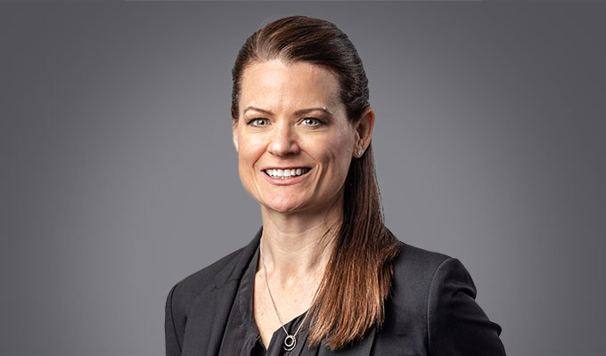 A woman with straight brown hair pulled back, wearing a black blazer and black top, smiles at the camera against a plain gray background.