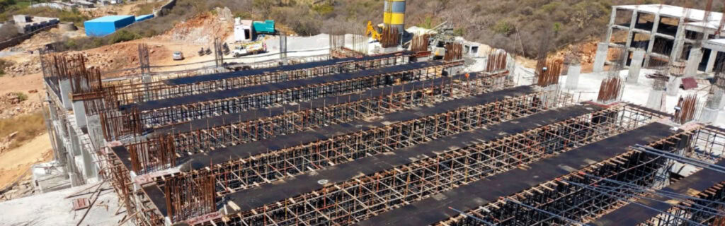 A construction site with multiple rows of rebar and formwork for concrete slabs, set amid unfinished buildings and machinery in a hilly area near Fairmont Palace, Udaipur.