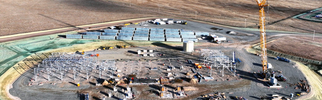 Aerial view of Cape Station construction site with steel framework being erected, a crane operating, building materials stacked nearby, and vehicles scattered around the area.