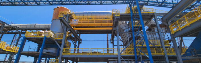 Large industrial facility with metal pipes, blue steel framework, and yellow railings under a clear blue sky—ideal for an iron ore briquetting plant.