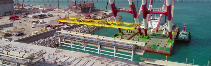 A large crane on a floating platform lifts a yellow steel structure at a coastal site, where precast concrete structures and breakwaters are visible among the buildings.