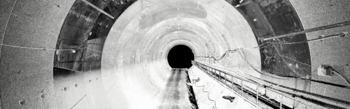 Black and white image of a long, empty tunnel—cables run along the right wall, hinting at digital technologies essential for modern infrastructure management. A dark circular opening appears at the far end.