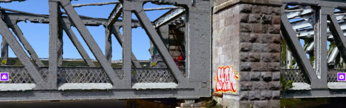 A close-up view of a metal railway bridge with stone supports and red and yellow graffiti on one pillar, ideal for an Asset Condition Survey or integration into a 3D Geospatial Platform.