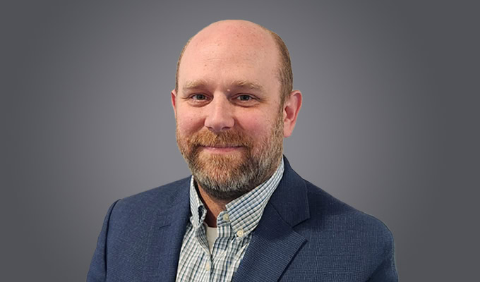 Brad Johnson, a man with a beard and bald head, wears a blue blazer and checked shirt as he poses in front of a plain grey background.