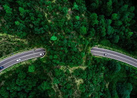Aerial view of two curved roads separated by dense green forest, with cars driving in opposite directions.