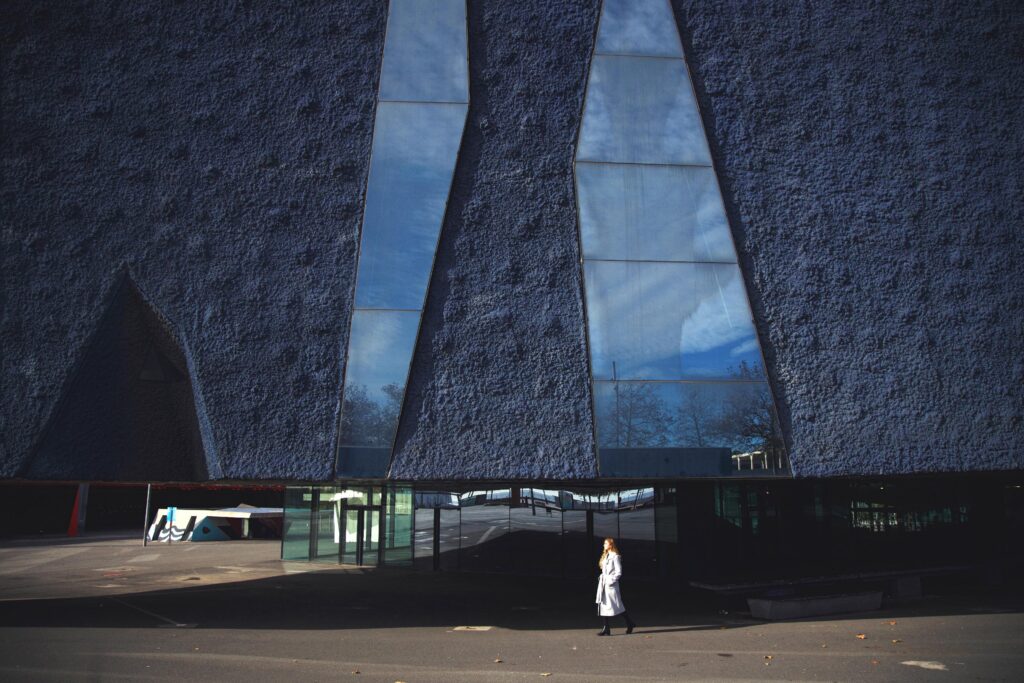 A person walks in front of a modern building with textured blue walls and tall, irregularly shaped reflective windows.