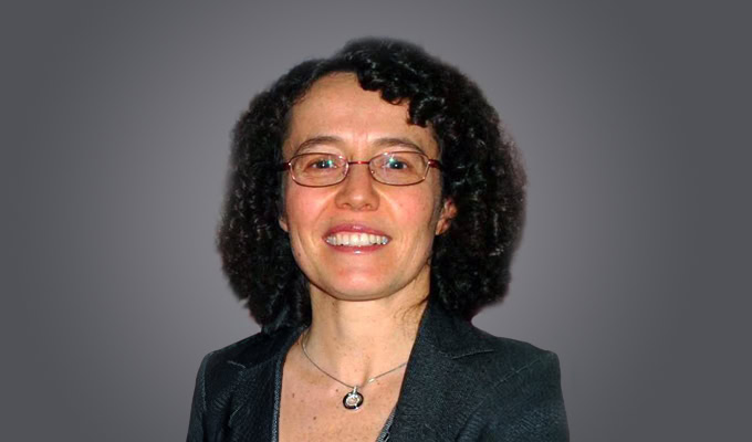 Catarina Freitas, a woman with curly dark hair, wearing glasses, a dark blazer, and a pendant necklace, smiles at the camera against a gray background.