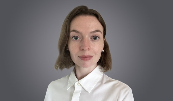 Woman with short brown hair wearing a white collared shirt, facing the camera with a neutral expression against a plain gray background.