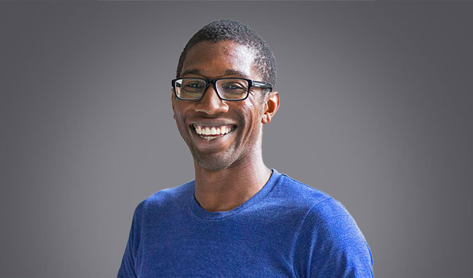 Christopher Jackson, wearing glasses and a blue t-shirt, smiles at the camera in front of a plain gray background.