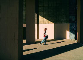 A person with red hair walks through a sunlit area between tall columns, casting long shadows on the ground and walls.