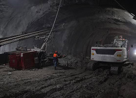 A construction worker in a safety vest stands near heavy machinery inside a dimly lit tunnel under construction.