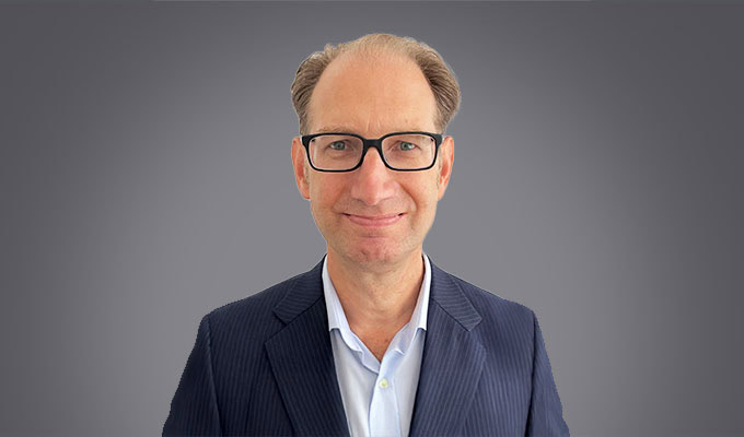 A middle-aged man with glasses, wearing a suit jacket and light blue shirt, stands in front of a plain grey background, facing the camera and smiling slightly.