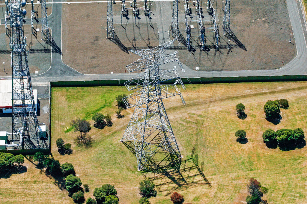 Aerial view of a tilted power transmission tower in a grassy area near an electrical substation, with scattered trees and visible shadows.