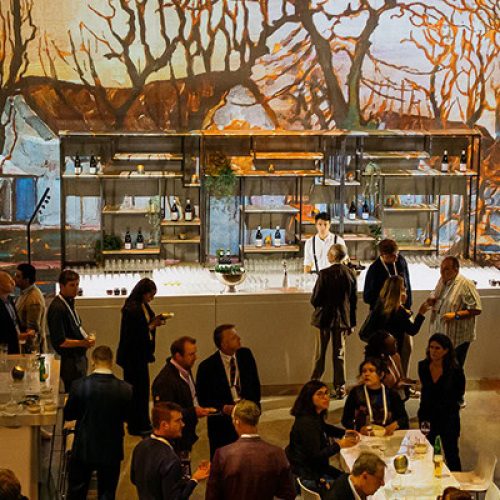 Large group of people in formal attire gather at an indoor event with a central bar and colorful mural of trees and fields on the walls.