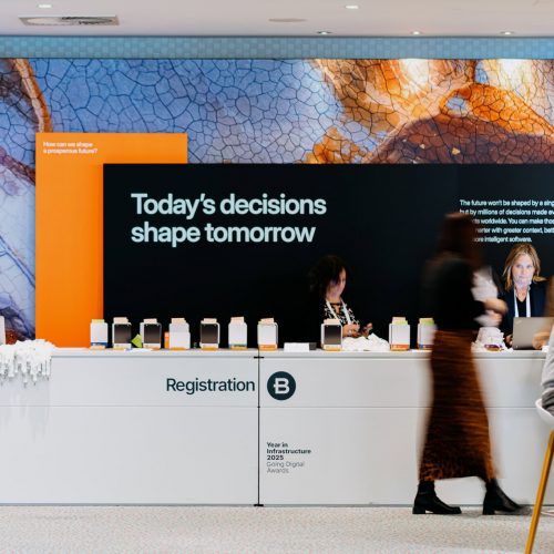 Registration desk at an event with staff assisting attendees; behind them, a large screen displays the text "Today's decisions shape tomorrow.