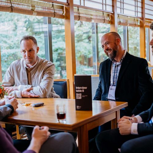 Four professionally dressed people sit around a wooden table in a well-lit room, engaged in conversation. A sign on the table reads “Interview table 8.”.
