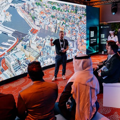 Man presenting in front of a wall-sized aerial-city display to a seated audience at a tech booth with iLab branding visible on the wall.