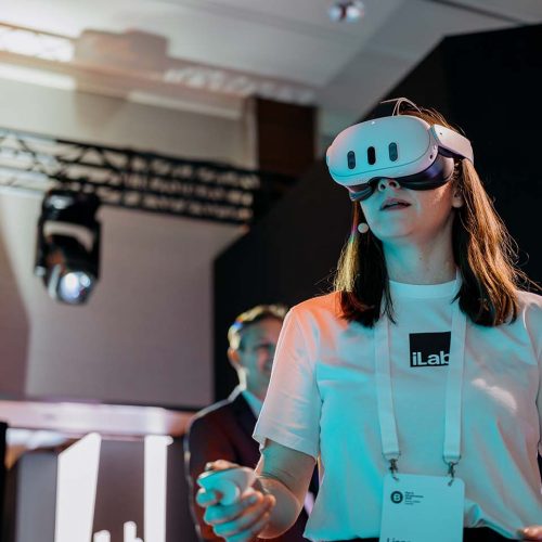 Woman wearing a VR headset at a technology demo, badge visible, reaching toward something off-screen in a conference setting
