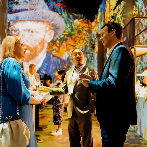 Group of professionals chatting at a colorful, art-filled reception with a buffet in the background far right and a standing woman in blue talking to a man in a suit.