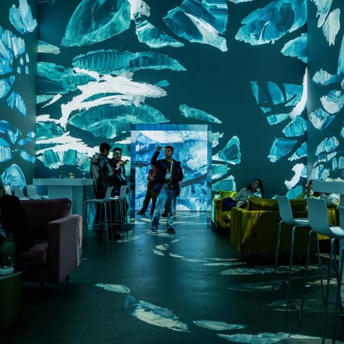 People mingle in a modern lounge with blue abstract wall projections and colorful seating areas.